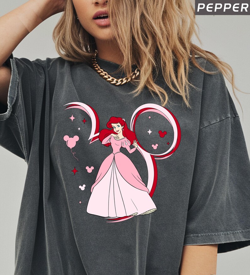 May include: Dark gray t-shirt featuring a cartoon illustration of Ariel from The Little Mermaid in a pink dress. The design includes a Mickey Mouse ear outline with the number 30, stars, and heart-shaped Mickey heads. The word "PEPPER" is at the top.