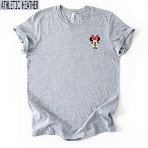 May include: An athletic heather gray short-sleeve t-shirt featuring a small graphic of Minnie Mouse on the upper left chest. The character is depicted with her signature red and white polka dot bow, white gloves, and a cheerful expression.