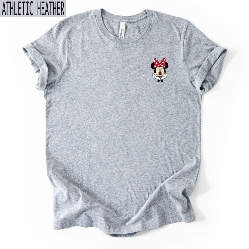 May include: An athletic heather gray short-sleeve t-shirt featuring a small graphic of Minnie Mouse on the upper left chest. The character is depicted with her signature red and white polka dot bow, white gloves, and a cheerful expression.