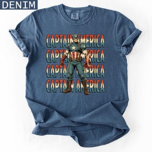 May include: Denim blue t-shirt featuring a Captain America graphic. The design includes the text "CAPTAIN AMERICA" in a stacked format, with a vintage comic book style illustration of Captain America holding his shield.