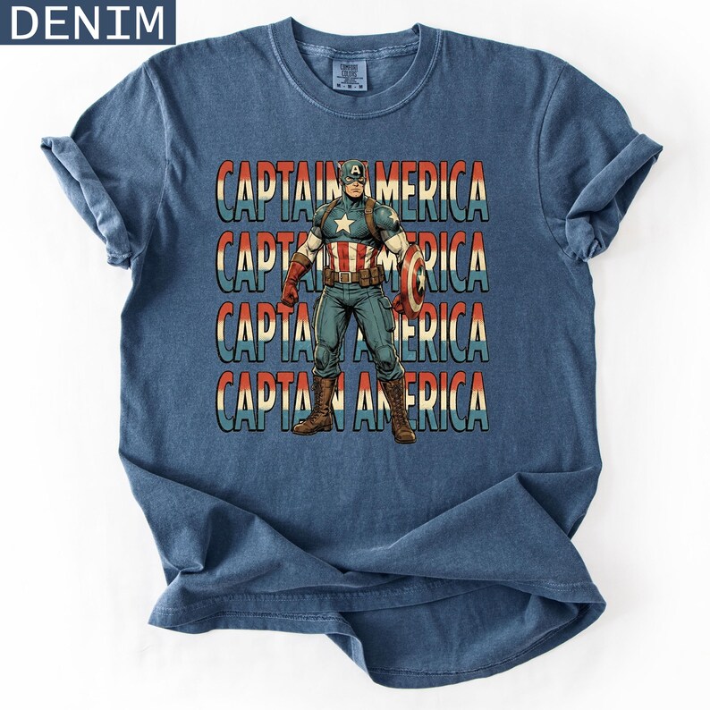 May include: Denim blue t-shirt featuring a Captain America graphic. The design includes the text "CAPTAIN AMERICA" in a stacked format, with a vintage comic book style illustration of Captain America holding his shield.