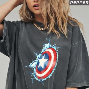 May include: Dark gray t-shirt featuring a Captain America shield graphic. The shield is red, white, and blue with a white star in the center, surrounded by a blue water splash effect. The word "PEPPER" is in the top right corner.