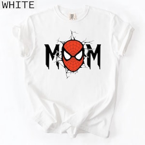 May include: A white short-sleeved t-shirt featuring a graphic print on the chest. The design displays the word MOM in black capital letters, with the letter O replaced by a red and black spider-web patterned mask with white eye lenses.