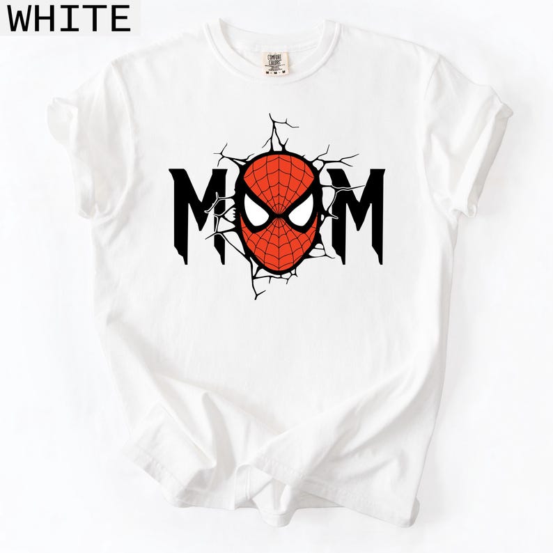 May include: A white short-sleeved t-shirt featuring a graphic print on the chest. The design displays the word MOM in black capital letters, with the letter O replaced by a red and black spider-web patterned mask with white eye lenses.