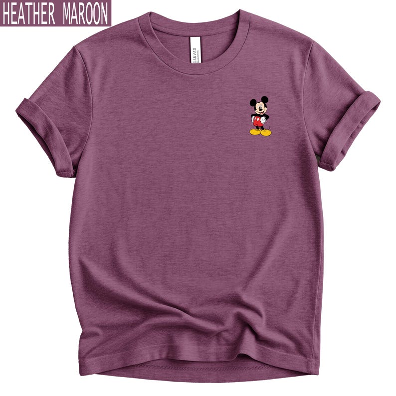 May include: A heather maroon short-sleeved t-shirt featuring a small graphic of Mickey Mouse on the upper left chest. The character is depicted with black ears, a white face, red shorts, and yellow shoes. The text HEATHER MAROON appears in the top left corner.