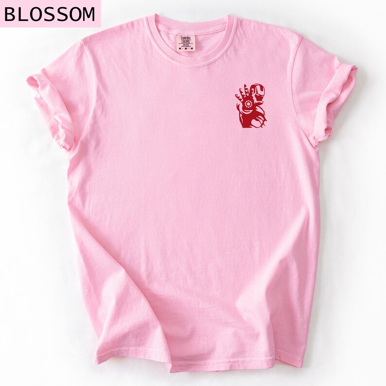 Puede incluir: Una camiseta de manga corta de color rosa claro con un peque&ntilde;o gr&aacute;fico de color rojo oscuro de un personaje rob&oacute;tico en el pecho izquierdo. El gr&aacute;fico muestra una figura con la mano extendida y un detalle circular. La palabra BLOSSOM est&aacute; impresa en negro.