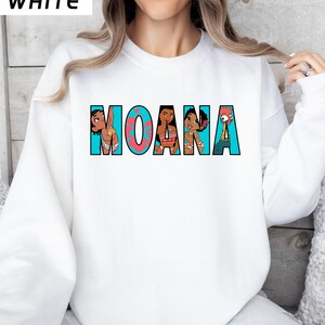 May include: White sweatshirt with the word "MOANA" in a colourful cartoon design. The letters are outlined in teal, with a coral-coloured "O". The design includes cartoon characters from the movie.