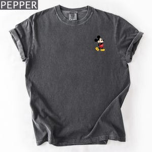 Pu&ograve; includere: T-shirt grigio scuro con una piccola grafica di Topolino sul davanti. Topolino indossa il suo abito classico: pantaloncini rossi, scarpe gialle e guanti bianchi. La maglietta ha le maniche arrotolate e la scritta "PEPPER" nell'angolo in alto a sinistra.