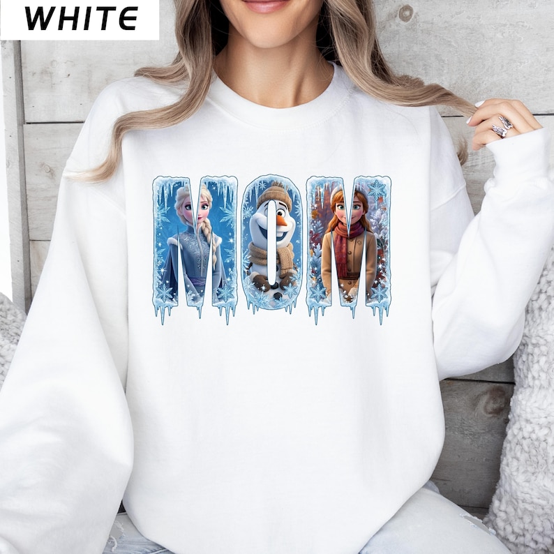 Puede incluir: Sudadera blanca con la palabra "MOM" en un dise&ntilde;o invernal, con personajes de una popular pel&iacute;cula de animaci&oacute;n. La palabra "WHITE" est&aacute; impresa en la esquina superior izquierda. El dise&ntilde;o incluye car&aacute;mbanos y copos de nieve.