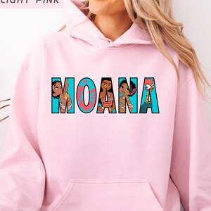 May include: Pale pink hoodie featuring the word "MOANA" in a colourful graphic design. The letters incorporate cartoon characters from the film. The hoodie has a front pocket and a hood.