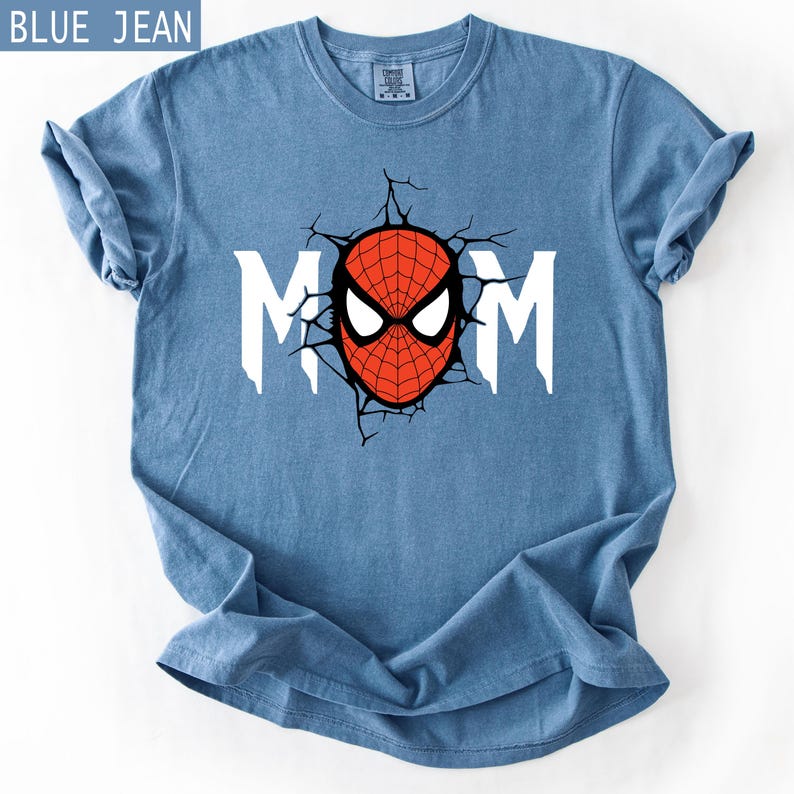 May include: A blue jean coloured short-sleeve t-shirt featuring a graphic print of the word MOM in white letters. The letter O is replaced by a red and black spider-themed mask design with black crack lines radiating outward from the centre.