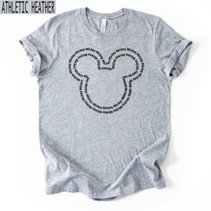 Peut inclure: T-shirt gris chin&eacute; avec un motif de t&ecirc;te de Mickey Mouse noir. La t&ecirc;te de Mickey Mouse est form&eacute;e par le mot "Mickey" r&eacute;p&eacute;t&eacute; en cercle. Le t-shirt est fait d'une mati&egrave;re douce et confortable.