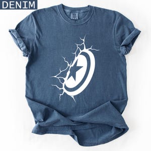 Pode incluir: Camiseta azul denim com um design gr&aacute;fico branco de escudo e estrela com efeito rachado. A palavra 'DENIM' est&aacute; no canto superior esquerdo. A camiseta est&aacute; dobrada sobre uma superf&iacute;cie branca.