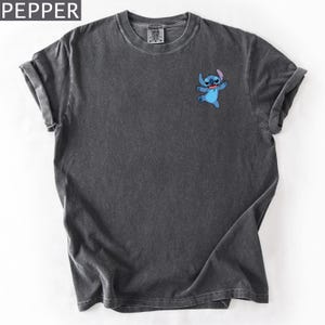 May include: Dark gray t-shirt featuring a small, embroidered image of the character Stitch from Lilo & Stitch. The shirt has a crew neck and rolled-up sleeves. The word "PEPPER" is in the top left corner.