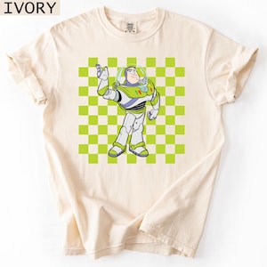 May include: Ivory-colored t-shirt featuring a cartoon illustration of Buzz Lightyear from Toy Story. The character is depicted in his signature space ranger suit, set against a green and white checkered background. The shirt has rolled-up sleeves.
