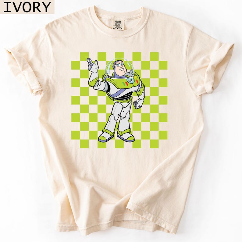 May include: Ivory-colored t-shirt featuring a cartoon illustration of Buzz Lightyear from Toy Story. The character is depicted in his signature space ranger suit, set against a green and white checkered background. The shirt has rolled-up sleeves.