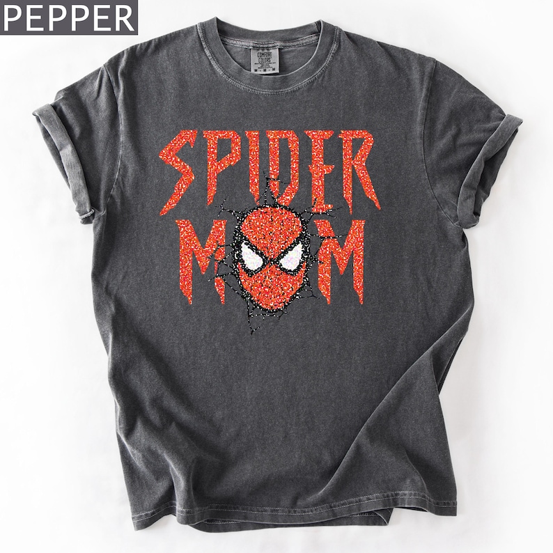 May include: Dark gray t-shirt with the words "SPIDER MOM" in red glitter lettering. The design includes a red and black spider mask graphic. The shirt is a crew neck with rolled-up sleeves.