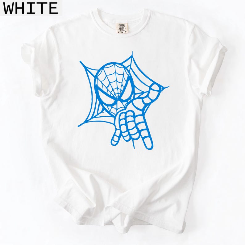 May include: White t-shirt with a blue graphic of a superhero's face and hand, surrounded by a spider web design. The word "WHITE" is printed in the upper left corner of the image.