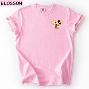 May include: A pale pink t-shirt with a small Mickey Mouse graphic. The cartoon mouse is depicted with his arms open. The word "BLOSSOM" is printed at the top left corner of the shirt.