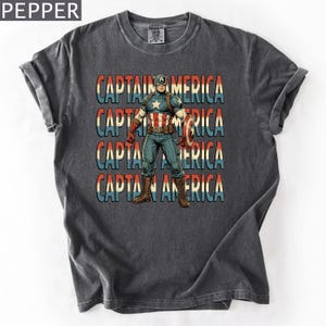 May include: Dark gray t-shirt featuring a Captain America graphic. The design includes the text "CAPTAIN AMERICA" in red, white, and blue, with an illustration of Captain America holding his shield. The shirt has a vintage, worn look.