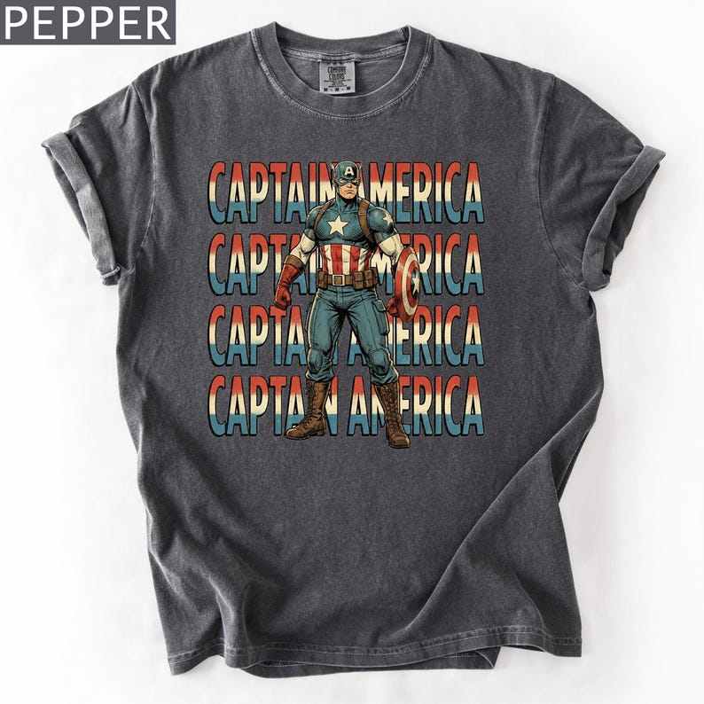 May include: Dark gray t-shirt featuring a Captain America graphic. The design includes the text "CAPTAIN AMERICA" in red, white, and blue, with an illustration of Captain America holding his shield. The shirt has a vintage, worn look.