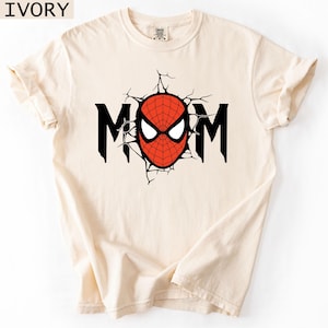 May include: An ivory short-sleeved t-shirt featuring a graphic print on the chest. The design displays the word MOM in black capital letters, with the letter O replaced by a red and black spider-web patterned mask with white eyes.