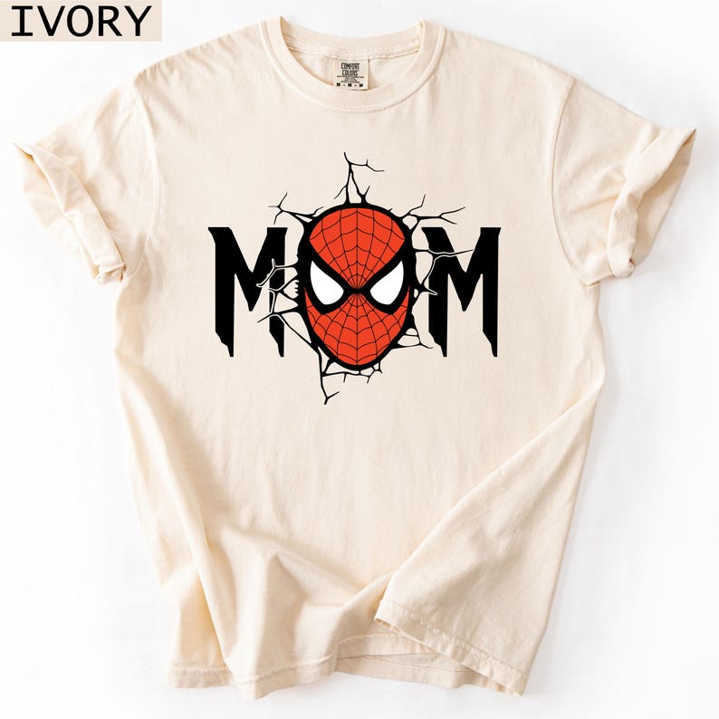May include: An ivory short-sleeved t-shirt featuring a graphic print on the chest. The design displays the word MOM in black capital letters, with the letter O replaced by a red and black spider-web patterned mask with white eyes.
