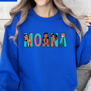 May include: Royal blue sweatshirt with the word "MOANA" in a colourful design. The letters are filled with cartoon images of characters from the film. The sweatshirt is a crew neck style.