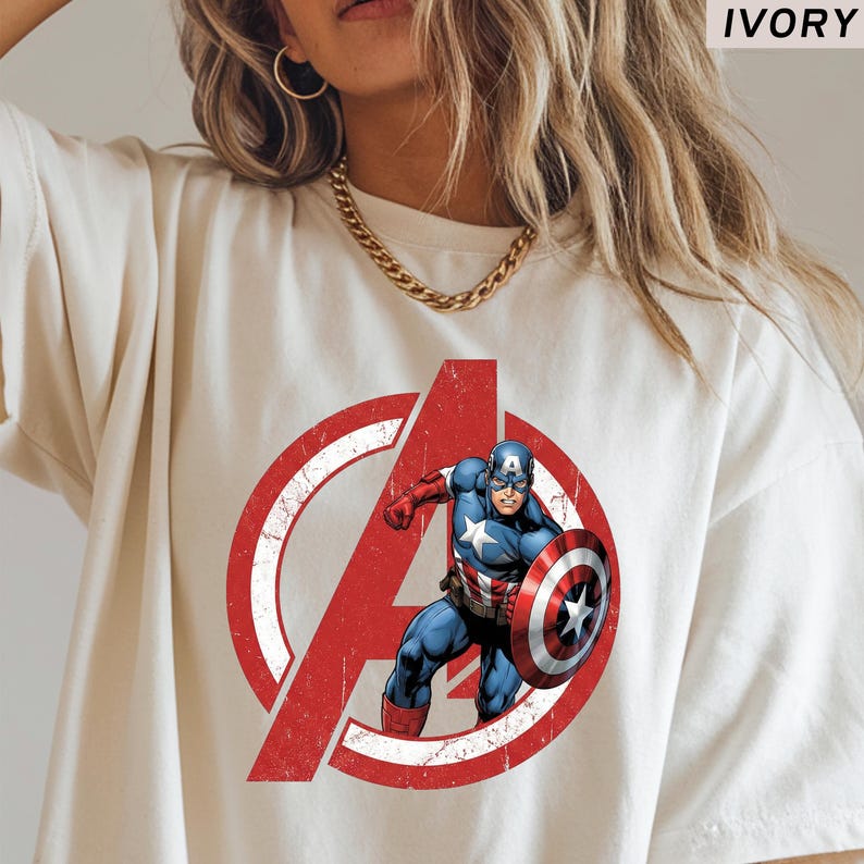 Puede incluir: Camiseta de color marfil con el logotipo desgastado de los Vengadores en rojo y blanco con el Capit&aacute;n Am&eacute;rica. El gr&aacute;fico del Capit&aacute;n Am&eacute;rica es azul, rojo y blanco, sosteniendo su escudo. La palabra "IVORY" est&aacute; impresa en la parte superior.