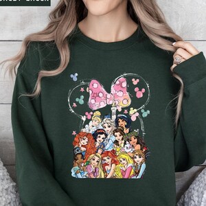 以下が含まれることがあります： ディズニーのプリンセスとピンクのドット柄のリボンが描かれた、フォレストグリーンのスウェットシャツ。デザインには、ミッキーマウスの耳の白いアウトラインとカラフルな蝶が含まれています。スウェットシャツは快適でカジュアルなスタイルです。