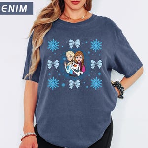 May include: Denim blue t-shirt featuring a graphic of characters from a popular animated film, surrounded by blue snowflakes and bows. The text "DENIM" is in a blue rectangle in the upper left corner. The shirt is a comfortable, casual style.