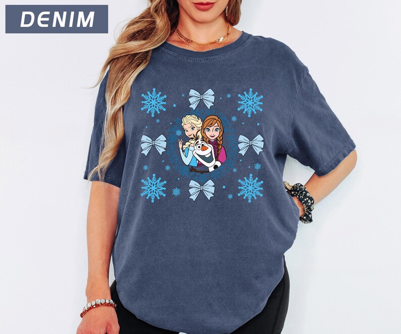 May include: Denim blue t-shirt featuring a graphic of characters from a popular animated film, surrounded by blue snowflakes and bows. The text "DENIM" is in a blue rectangle in the upper left corner. The shirt is a comfortable, casual style.