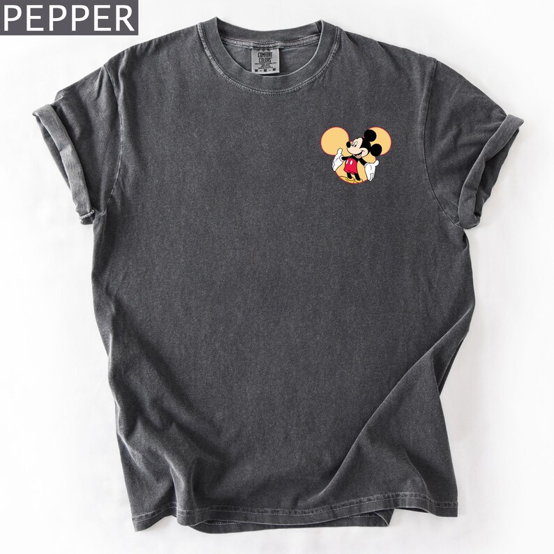 May include: Dark grey t-shirt with a small Mickey Mouse graphic on the chest. The graphic shows Mickey Mouse with his arms out, set against a yellow circle. The shirt has rolled-up sleeves and a crew neck.