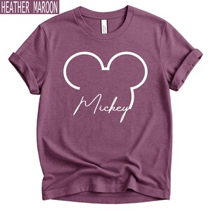 May include: Heather maroon t-shirt featuring a white Mickey Mouse outline and the name "Mickey" in a script font. The shirt has rolled-up sleeves and a crew neck. The text "Heather Maroon" is in the top left corner.