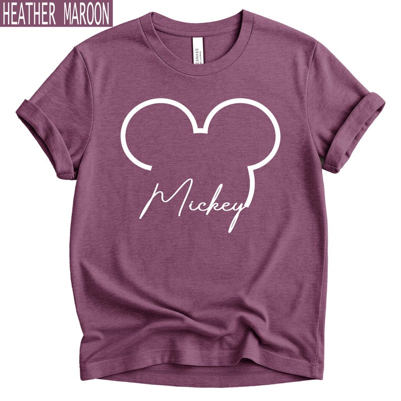May include: Heather maroon t-shirt featuring a white Mickey Mouse outline and the name "Mickey" in a script font. The shirt has rolled-up sleeves and a crew neck. The text "Heather Maroon" is in the top left corner.