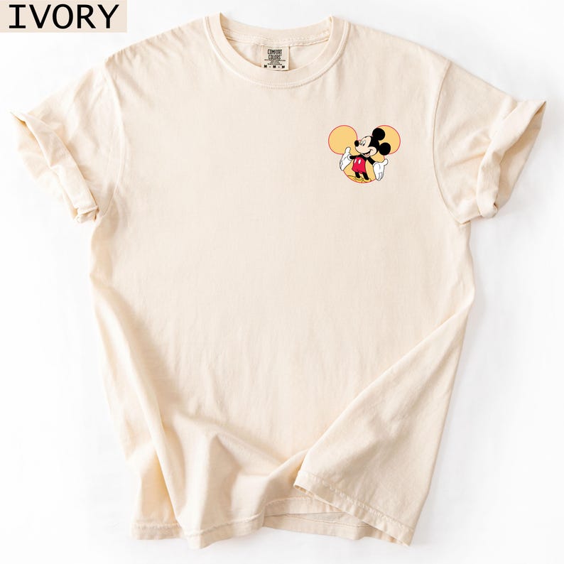 May include: Ivory-coloured t-shirt with a small Mickey Mouse graphic. The cartoon character is shown in his classic pose, with yellow ears, a red shirt, and white gloves. The shirt has short sleeves and a crew neck. The word "IVORY" is in the top left corner.