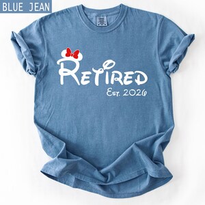 Puede incluir: Camiseta azul vaquero con la palabra "Retired" en escritura blanca, con un lazo de Minnie Mouse. El texto "Est. 2026" est&aacute; debajo de la palabra "Retired". La camiseta est&aacute; doblada y tiene las mangas enrolladas.