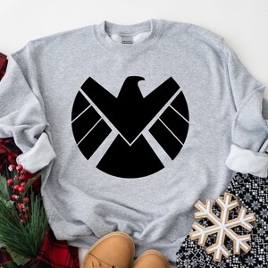 Puede incluir: Sudadera gris con un logotipo negro de S.H.I.E.L.D. impreso en la parte delantera.