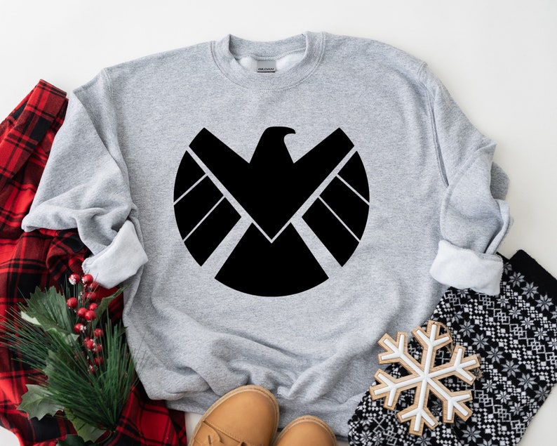 Puede incluir: Sudadera gris con un logotipo negro de S.H.I.E.L.D. impreso en la parte delantera.