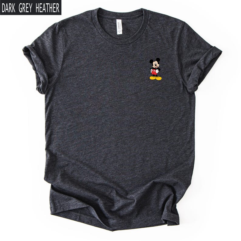 May include: A dark grey heather crew neck t-shirt featuring a small graphic of Mickey Mouse on the left chest. The character is depicted with black ears, a red pair of shorts, and yellow shoes. The text "DARK GREY HEATHER" appears in the top left corner.
