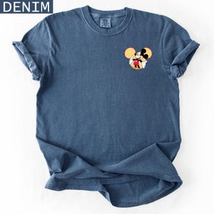 May include: Denim blue t-shirt featuring a small Mickey Mouse graphic on the chest. The graphic shows Mickey Mouse in his classic pose, with yellow ears and a red and black outfit. The shirt has rolled-up sleeves.