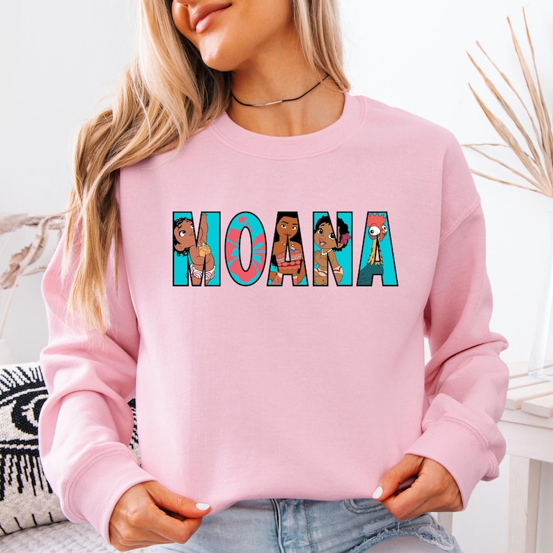 May include: A pale pink sweatshirt with the word "MOANA" printed across the front in a turquoise and black font. The letters are filled with cartoon images of characters from the film. The sweatshirt has a crew neck and long sleeves.