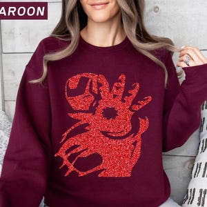 May include: Maroon sweatshirt with a red glitter graphic design. The graphic is a stylized reindeer head. The word "MAROON" is in a white font in the upper left corner of the image. The sweatshirt is a solid maroon color.