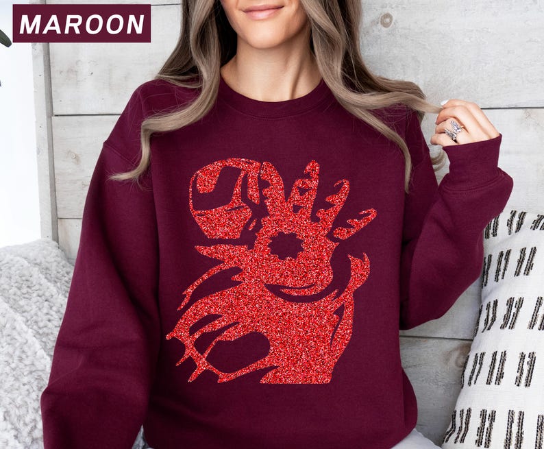 May include: Maroon sweatshirt with a red glitter graphic design. The graphic is a stylized reindeer head. The word "MAROON" is in a white font in the upper left corner of the image. The sweatshirt is a solid maroon color.