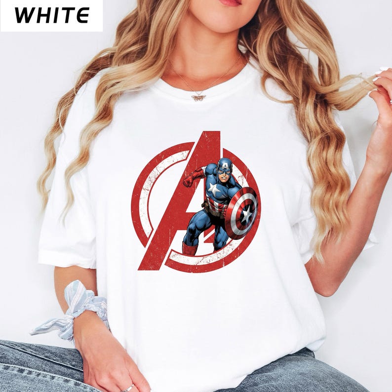 Puede incluir: Camiseta blanca con el logotipo rojo desgastado de los Vengadores y el Capit&aacute;n Am&eacute;rica. El superh&eacute;roe est&aacute; representado con su traje azul, rojo y blanco, sosteniendo su escudo. La palabra "WHITE" est&aacute; impresa arriba a la izquierda.