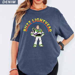 Op de afbeelding: Een denim blauw T-shirt met een afbeelding van Buzz Lightyear uit Toy Story. Het personage staat met gekruiste armen en de woorden "BUZZ LIGHTYEAR" staan er in een boog boven in geel, rood en groen.
