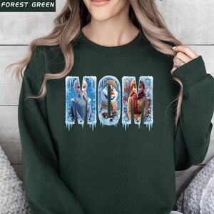 Puede incluir: Sudadera verde oscuro con la palabra "MOM" en un dise&ntilde;o de invierno. Las letras presentan personajes de la pel&iacute;cula Frozen, incluyendo a Elsa, Olaf y Anna, sobre un fondo de hielo y copos de nieve.