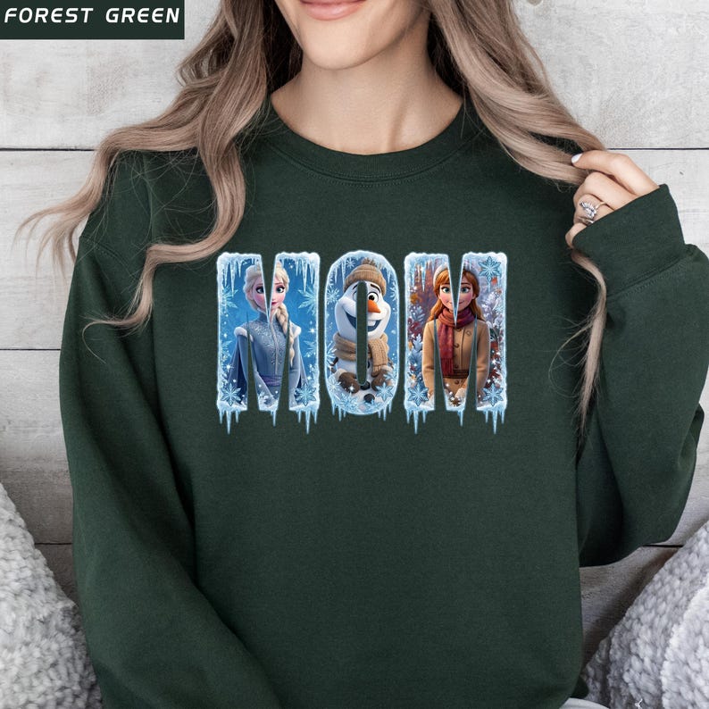 Puede incluir: Sudadera verde oscuro con la palabra "MOM" en un dise&ntilde;o de invierno. Las letras presentan personajes de la pel&iacute;cula Frozen, incluyendo a Elsa, Olaf y Anna, sobre un fondo de hielo y copos de nieve.