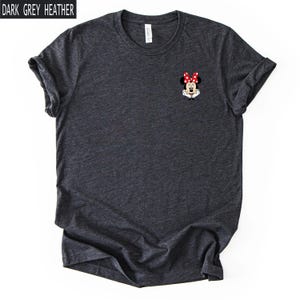 May include: A dark grey heather crew neck t-shirt featuring a small graphic of Minnie Mouse on the left chest. The character is depicted with her signature red and white polka dot bow, white gloves, and black ears against the dark fabric background.