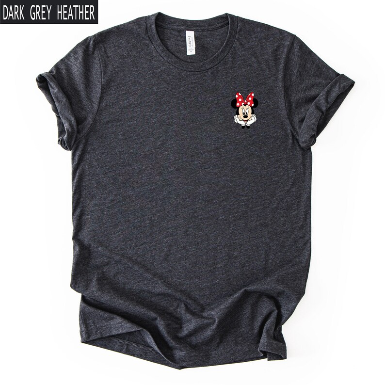 May include: A dark grey heather crew neck t-shirt featuring a small graphic of Minnie Mouse on the left chest. The character is depicted with her signature red and white polka dot bow, white gloves, and black ears against the dark fabric background.